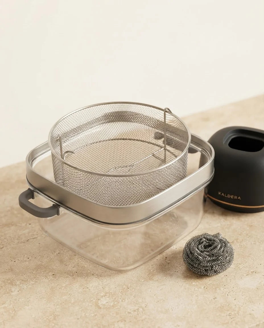 Close-up of the bare stainless steel mesh basket of Kaldera Glass resting in the clear glass bowl, a scouring pad beside it